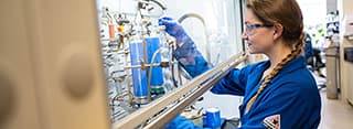 A woman with a side braid in a blue lab coat, blue lab gloves and safety glasses works conducts research in a lab