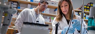 A man with a lab coat and safety glasses, and a woman in a lab coat with gloves, doing research in a lab