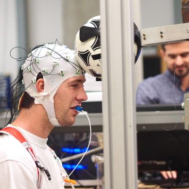 A person wearing a sensor-covered cap is performing an experiment involving a soccer ball, with two people observing in the background.